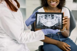 Close up cropped image of female dentist and african american woman patient at dentist's office, holding together digital tablet with xray panoramic jaw and teeth image. Oral care and check up